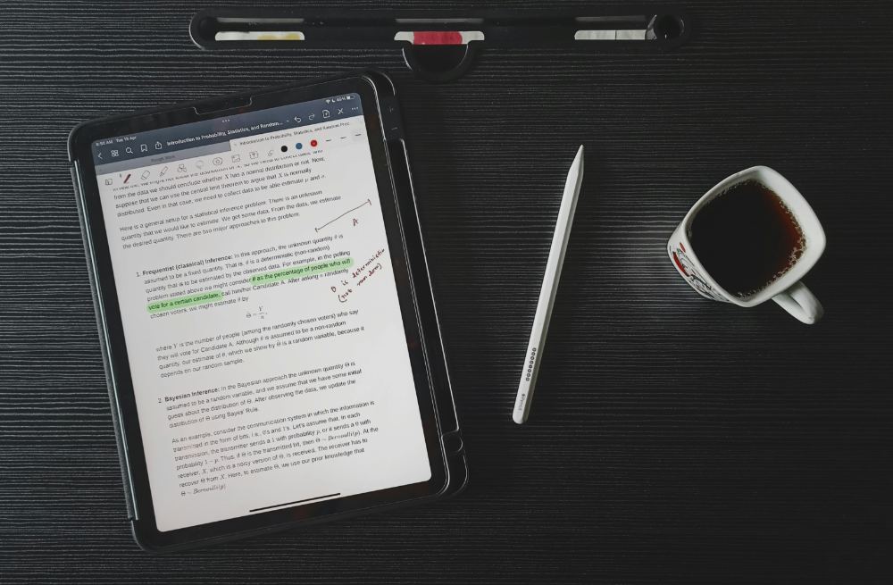A document open on a tablet, marked up in green highlighter and red pen, beside a stylus and a cup of coffee.